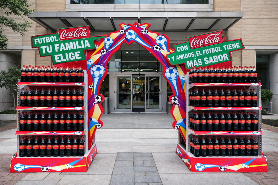 Exhibidor POP de Coca-Cola con arco temático de fútbol en entrada de edificio.